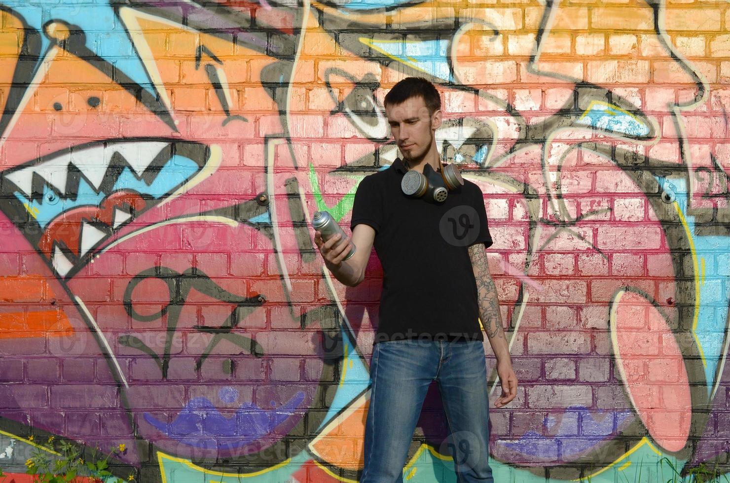 Young caucasian graffiti artist in black t-shirt with silver aerosol spray can near colorful graffiti in pink tones on brick wall. Street art and contemporary painting process photo