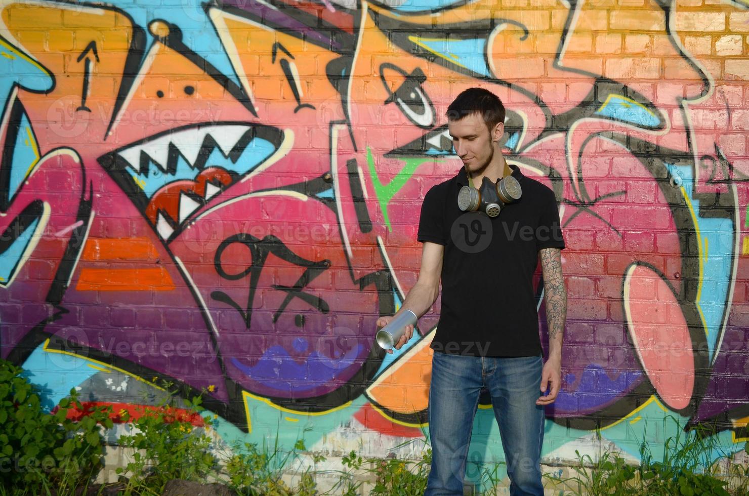 Young caucasian graffiti artist in black t-shirt with silver aerosol spray can near colorful graffiti in pink tones on brick wall. Street art and contemporary painting process photo