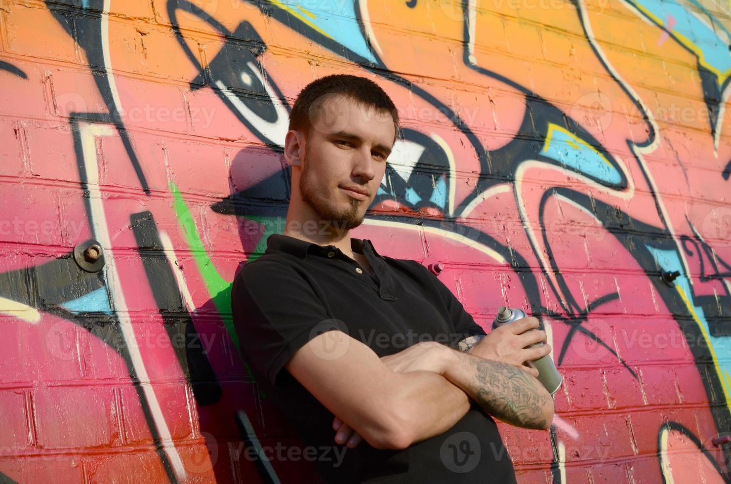 Young caucasian graffiti artist in black t-shirt with silver aerosol spray can near colorful graffiti in pink tones on brick wall. Street art and contemporary painting process photo