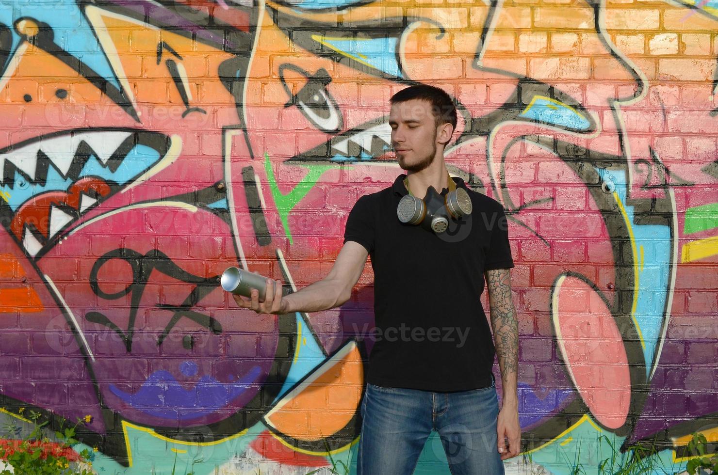 Young caucasian graffiti artist in black t-shirt with silver aerosol spray can near colorful graffiti in pink tones on brick wall. Street art and contemporary painting process photo