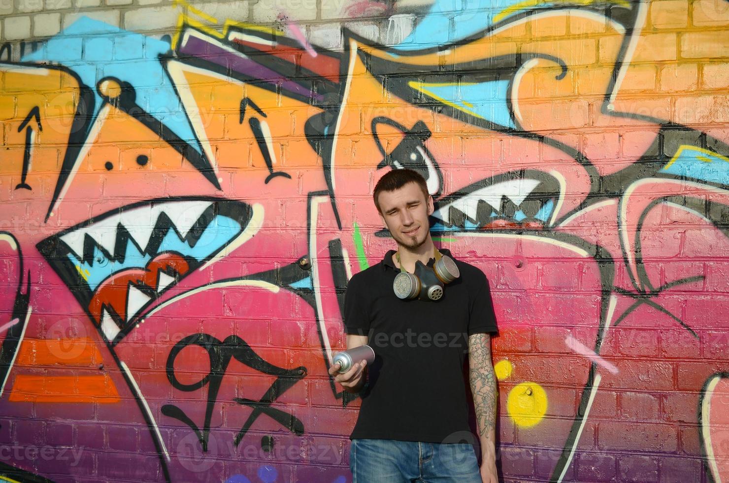 Young caucasian graffiti artist in black t-shirt with silver aerosol spray can near colorful graffiti in pink tones on brick wall. Street art and contemporary painting process photo