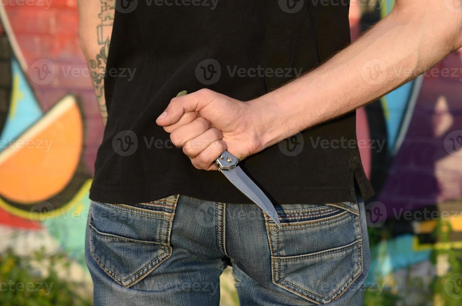 vista trasera de un joven caucásico con un cuchillo en la mano contra la pared de ladrillo del ghetto con pinturas de graffiti. concepto de fuerzas criminales y cargo de agresión foto