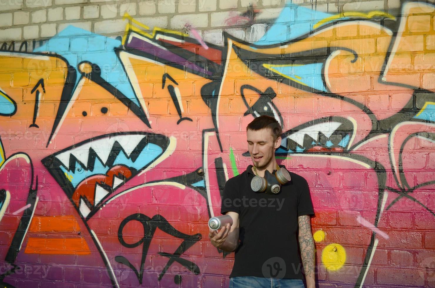 Young caucasian graffiti artist in black t-shirt with silver aerosol spray can near colorful graffiti in pink tones on brick wall. Street art and contemporary painting process photo