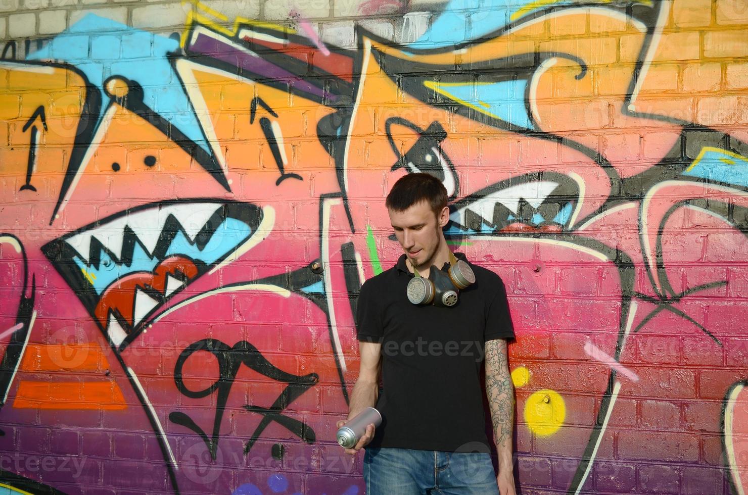 Young caucasian graffiti artist in black t-shirt with silver aerosol spray can near colorful graffiti in pink tones on brick wall. Street art and contemporary painting process photo