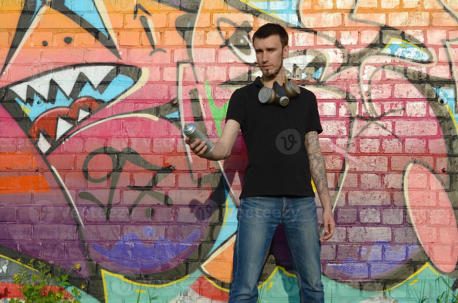 Young caucasian graffiti artist in black t-shirt with silver aerosol spray can near colorful graffiti in pink tones on brick wall. Street art and contemporary painting process photo