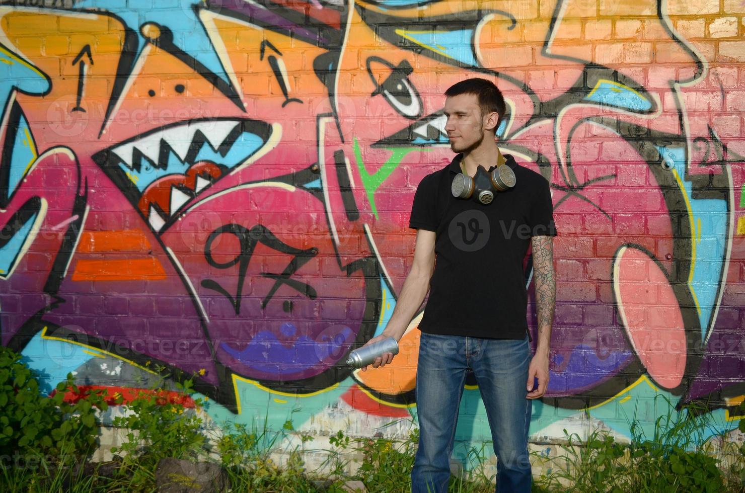 Young caucasian graffiti artist in black t-shirt with silver aerosol spray can near colorful graffiti in pink tones on brick wall. Street art and contemporary painting process photo