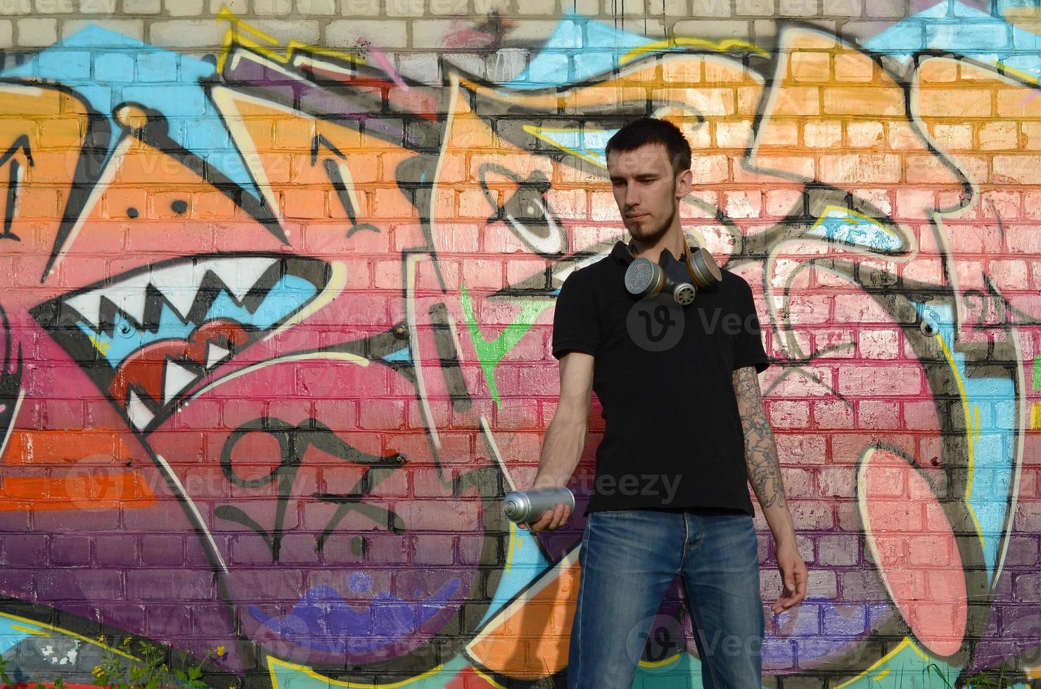 Young caucasian graffiti artist in black t-shirt with silver aerosol spray can near colorful graffiti in pink tones on brick wall. Street art and contemporary painting process photo