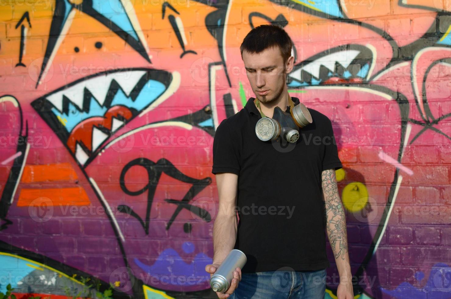 Young caucasian graffiti artist in black t-shirt with silver aerosol spray can near colorful graffiti in pink tones on brick wall. Street art and contemporary painting process photo