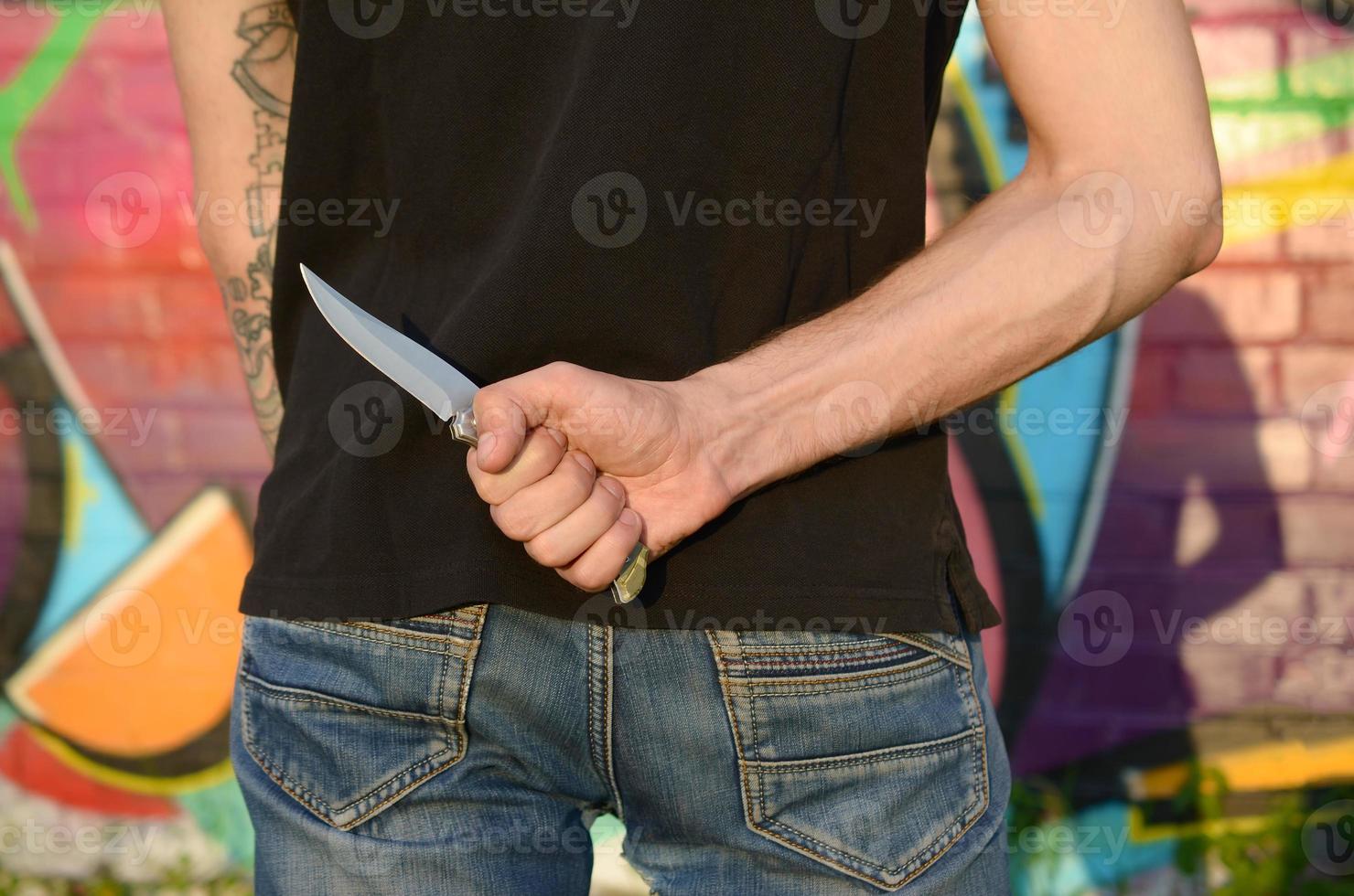 Back view of young caucasian man with knife in his hand against ghetto brick wall with graffiti paintings. Concept of criminal forces and aggression charge photo