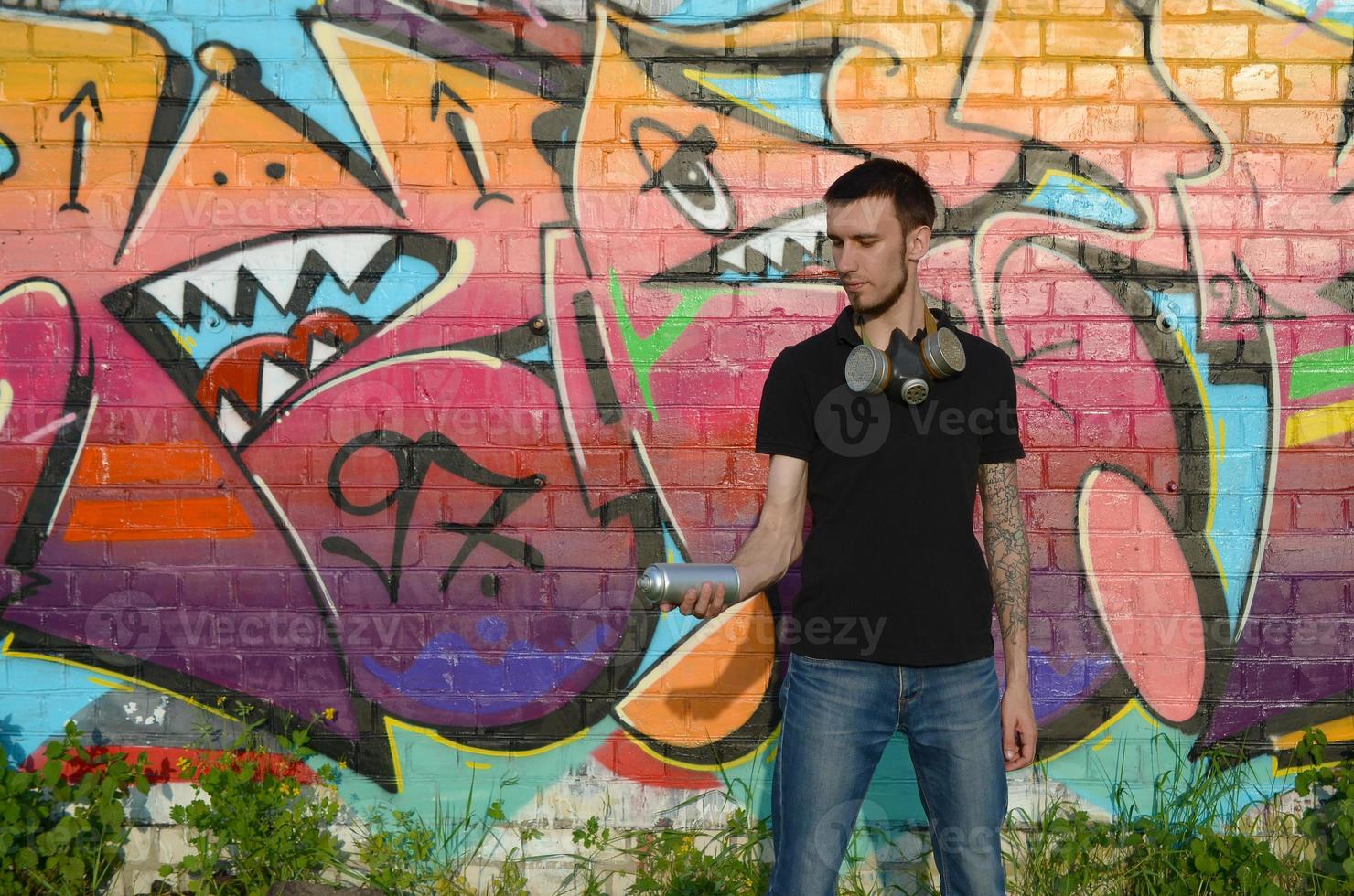 Young caucasian graffiti artist in black t-shirt with silver aerosol spray can near colorful graffiti in pink tones on brick wall. Street art and contemporary painting process photo