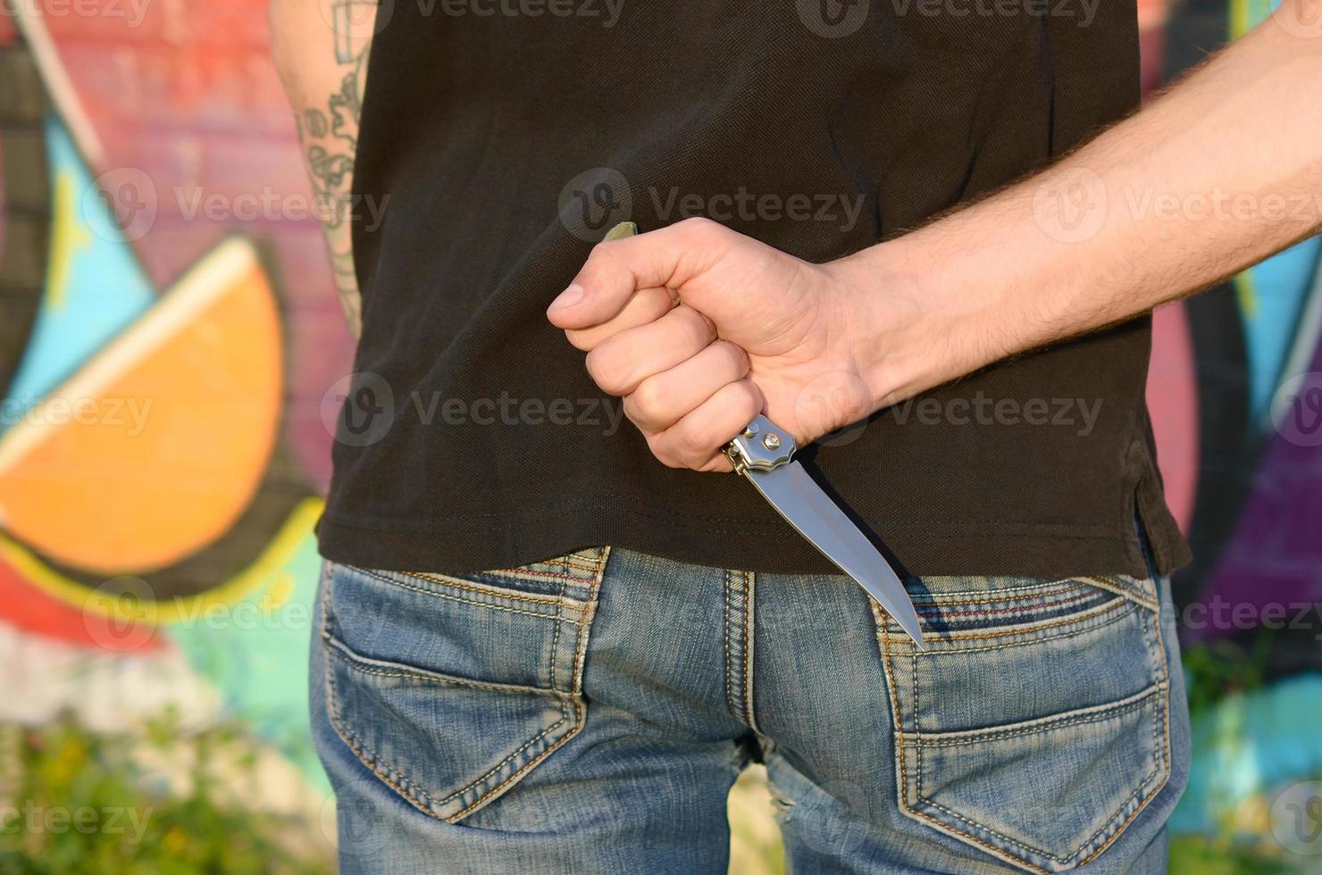 Back view of young caucasian man with knife in his hand against ghetto brick wall with graffiti paintings. Concept of criminal forces and aggression charge photo