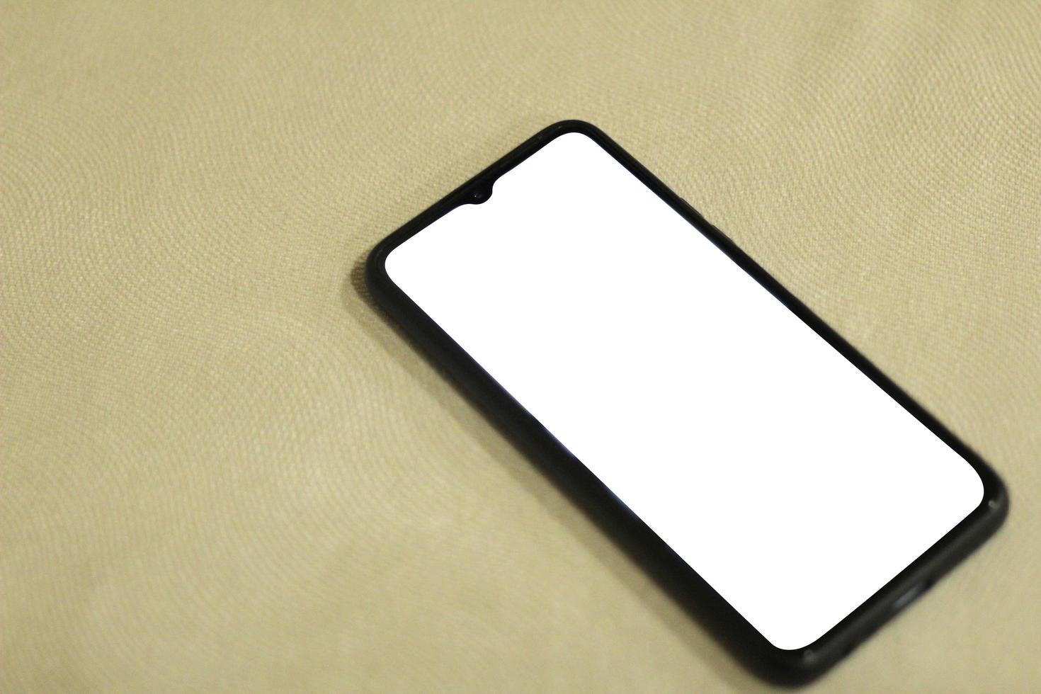 A black cell phone with a blank white screen sits on a bench upholstered in a textured cloth. Suitable for cellphone or gadget mockups photo