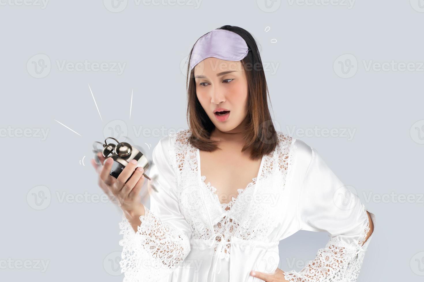 Asian woman in a white satin nightgown and wearing lace robe shocked as he wakes up late. A girl who wakes up late looks at the alarm clock on a light gray background. photo