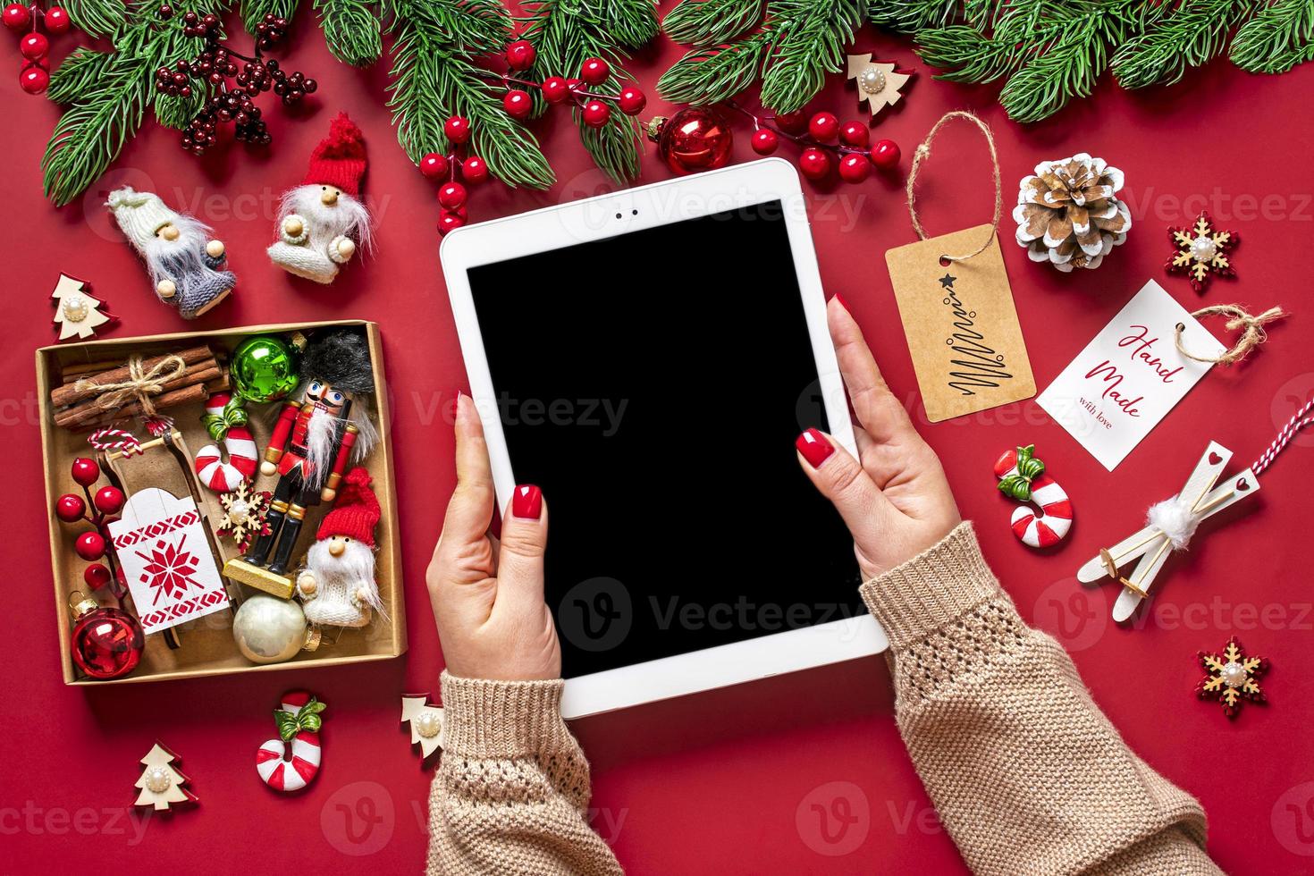 top view female hands holding tablet with black digital screen, Christmas box and decor on red table Flat lay Holiday shopping list, Happy New Year, online shop, chooses gifts, makes purchases Mockup photo