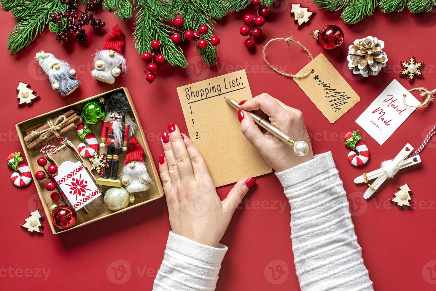female hand holds pen and writing Christmas shopping list, gift ideas on white notepad on red background Top view Flat lay Holiday card Woman making Christmas presents list, Planning, buying concept photo