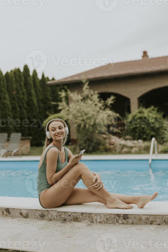 Young woman sitting by the swimming pool and listen music from mobile phone with headphones in the house backyard photo