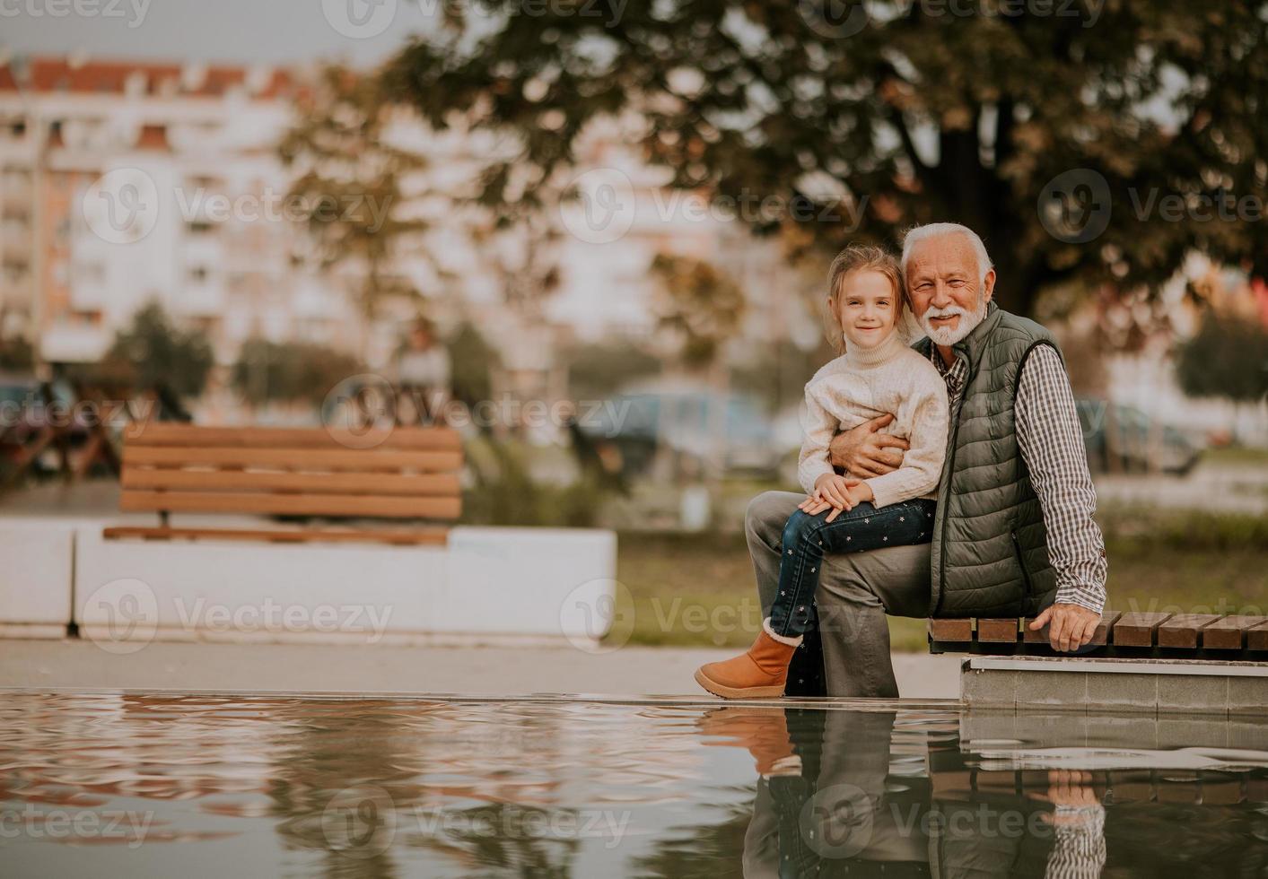 Grandfather spending time with his granddaughter by small water pool in park on autumn day photo