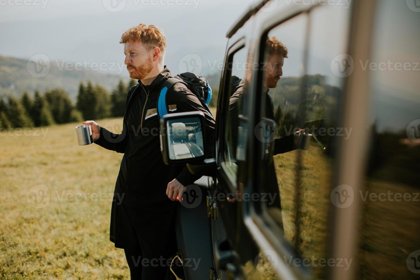 Young man relaxing and drinking coffee by the terrain vehicle hood at countryside photo