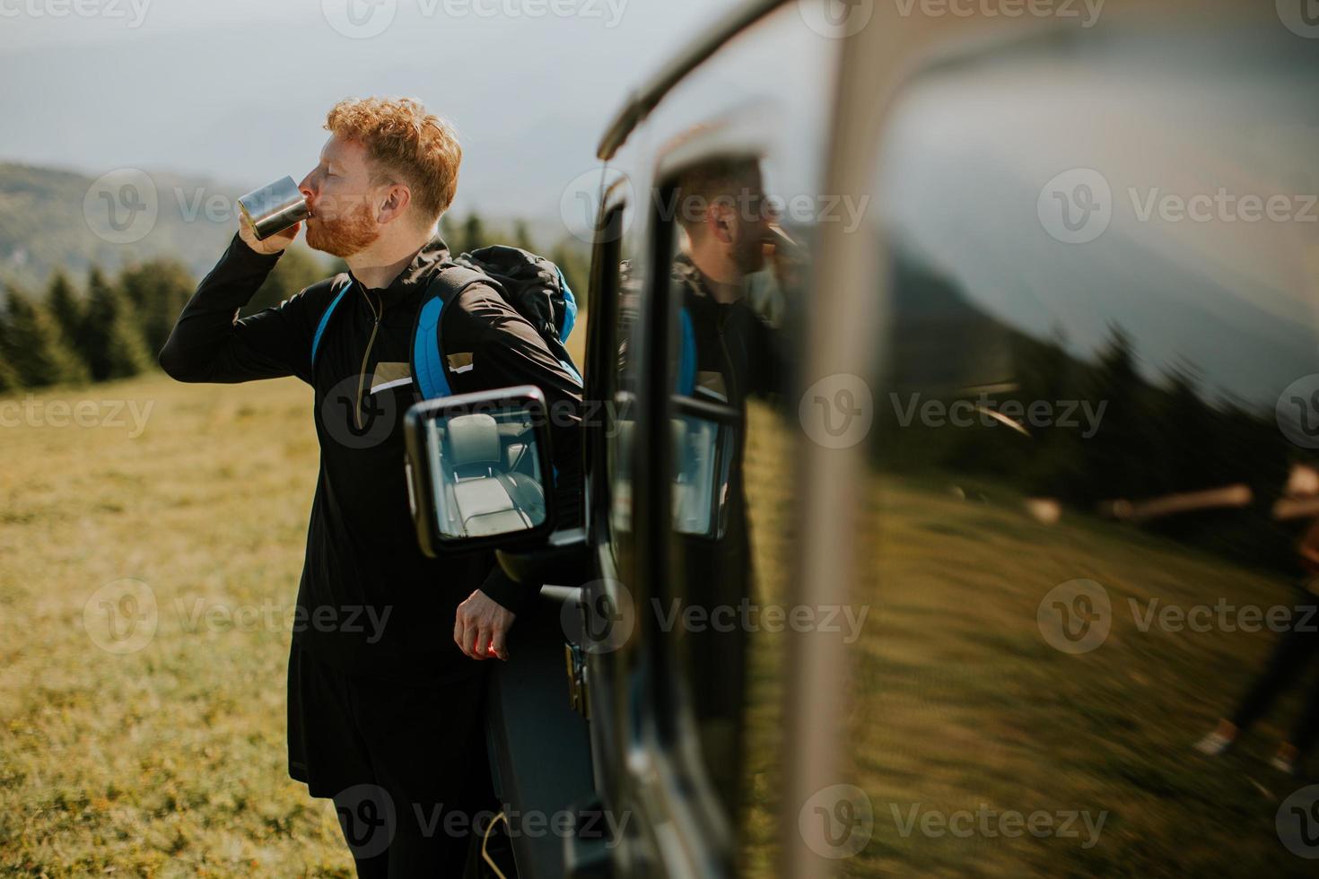 Young man relaxing and drinking coffee by the terrain vehicle hood at countryside photo