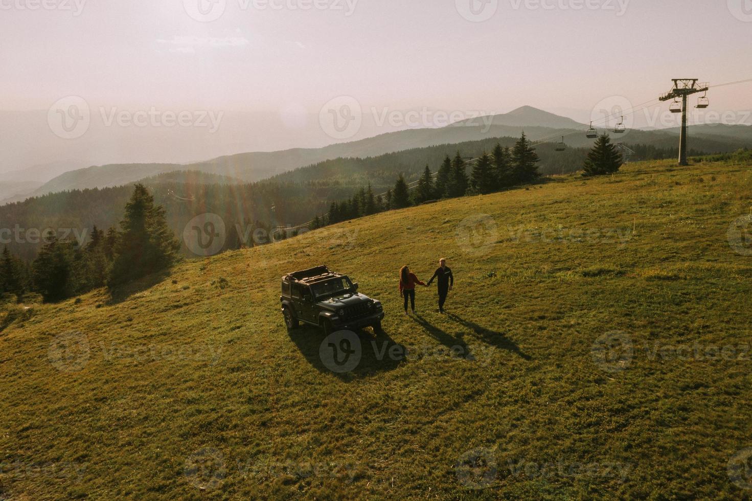 Aerial view at couple walking with backpacks by terrain vehicle over green hills photo