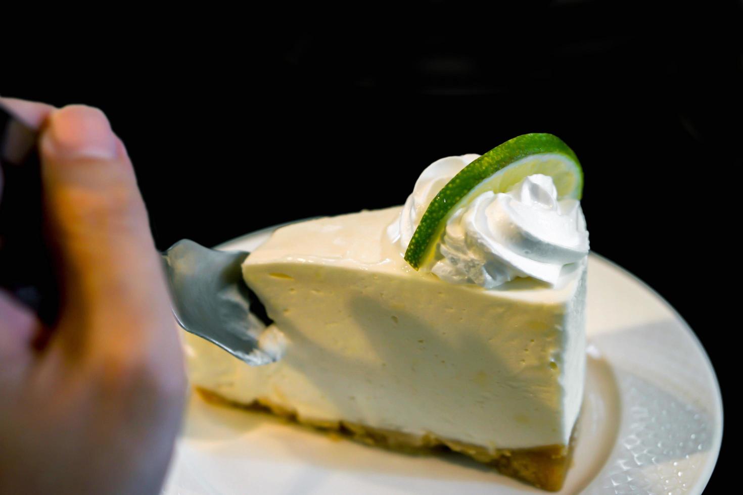 Image of hand holding a teaspoon is dividing a cake from a lime cream cake on a white ceramic plate and black background. photo