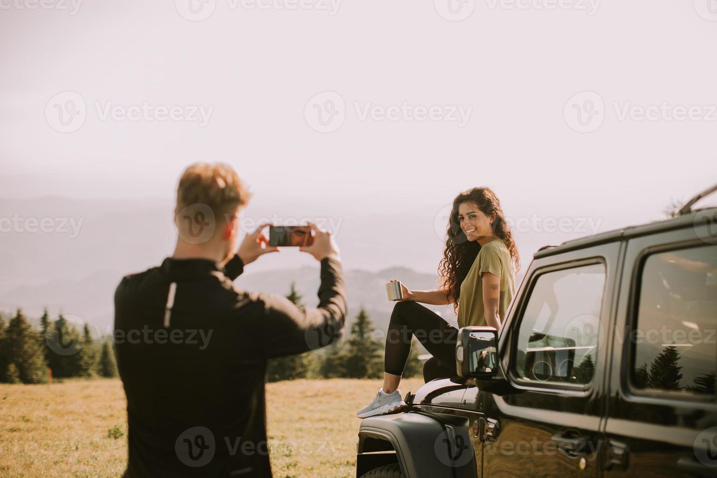 Young couple relaxing on a terrain vehicle hood and taking photo with mobile phone at countryside
