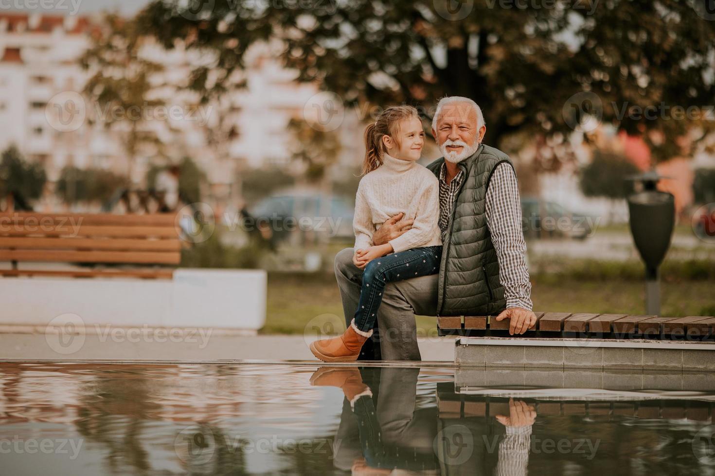 el abuelo pasa tiempo con su nieta junto a una pequeña piscina de agua en el parque el día de otoño foto