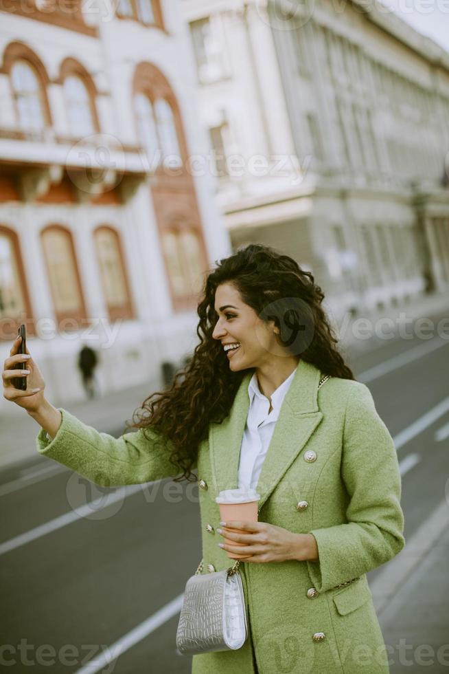 mujer joven que usa un teléfono inteligente para tomar una foto selfie en la calle y toma café para llevar