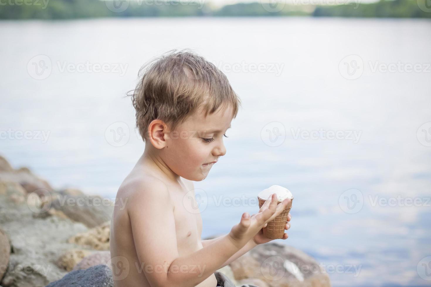 A cute blond boy appetizingly eats ice cream in the summer, sitting on the bank of the river. Cool off by the water. Funny facial expression. photo