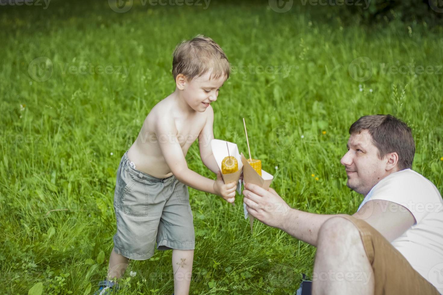 A cute blond boy and his father eat corn in the summer, sitting on the bank of the river on the juicy grass. Funny facial expression. photo