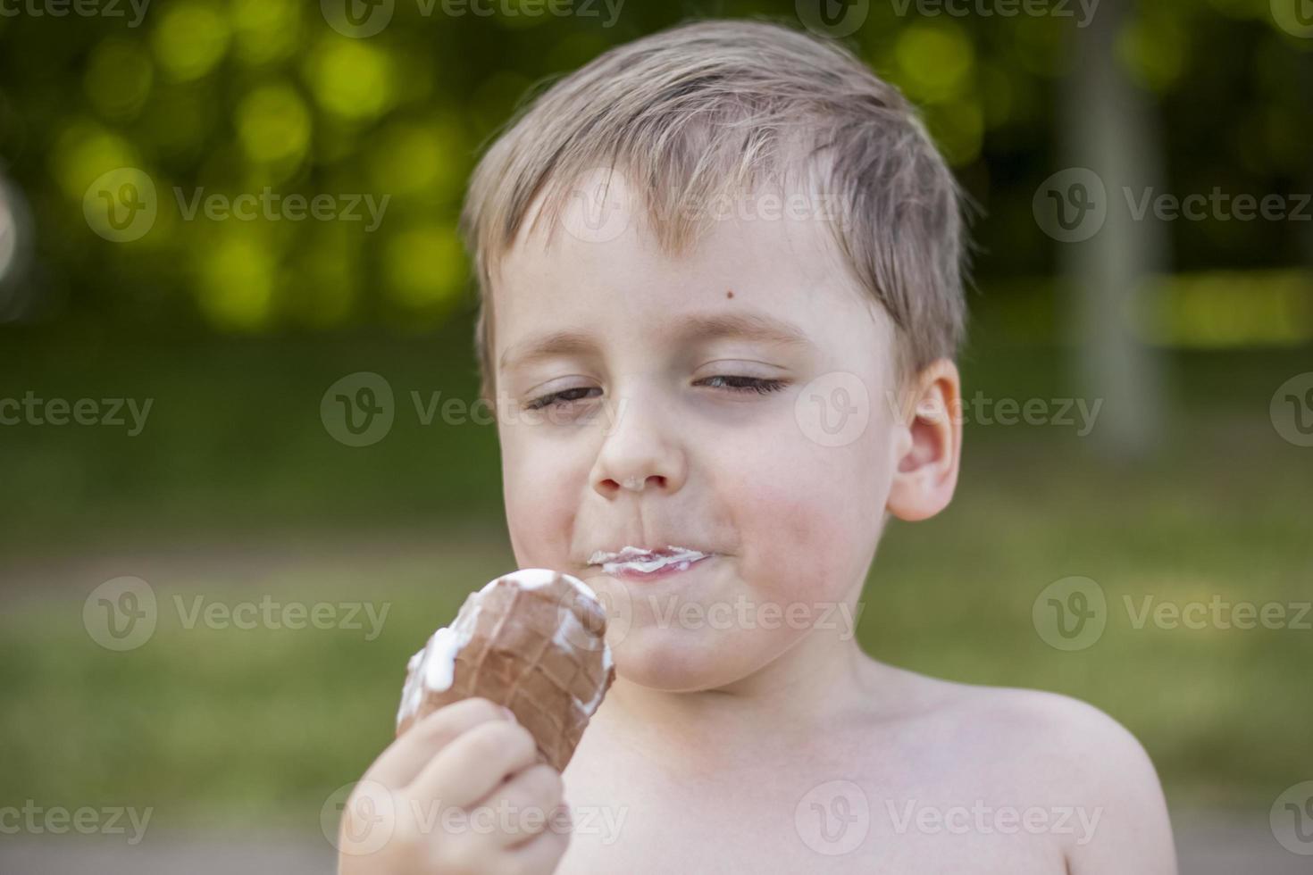 A cute blond boy appetizingly eats ice cream in the summer, sitting on the bank of the river. Cool off by the water. Funny facial expression. photo