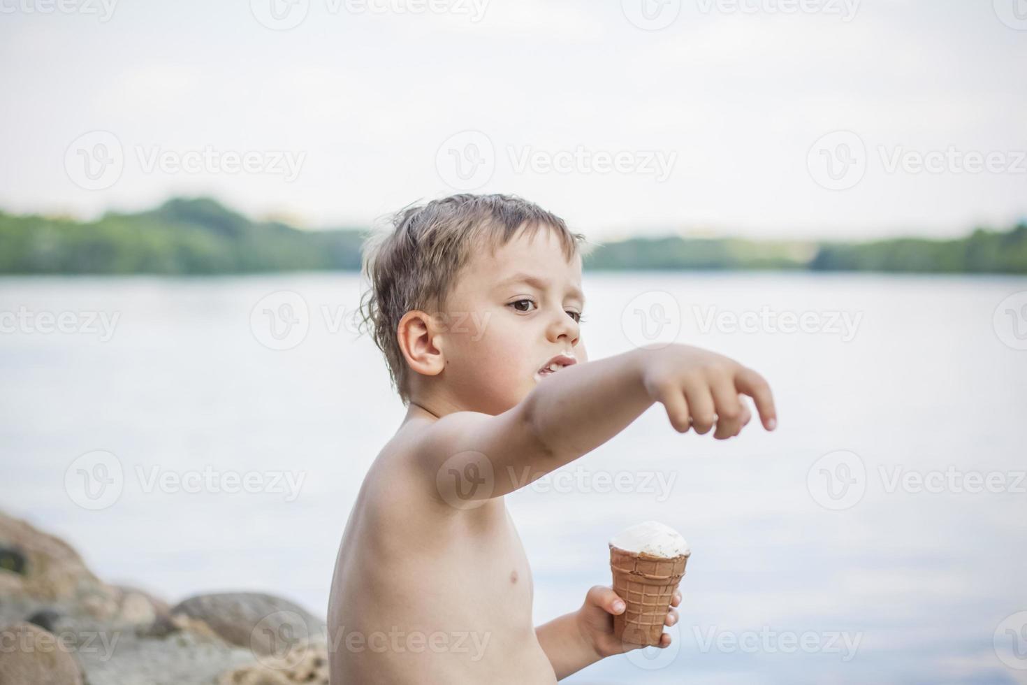 A cute blond boy appetizingly eats ice cream in the summer, sitting on the bank of the river. Cool off by the water. Funny facial expression. photo
