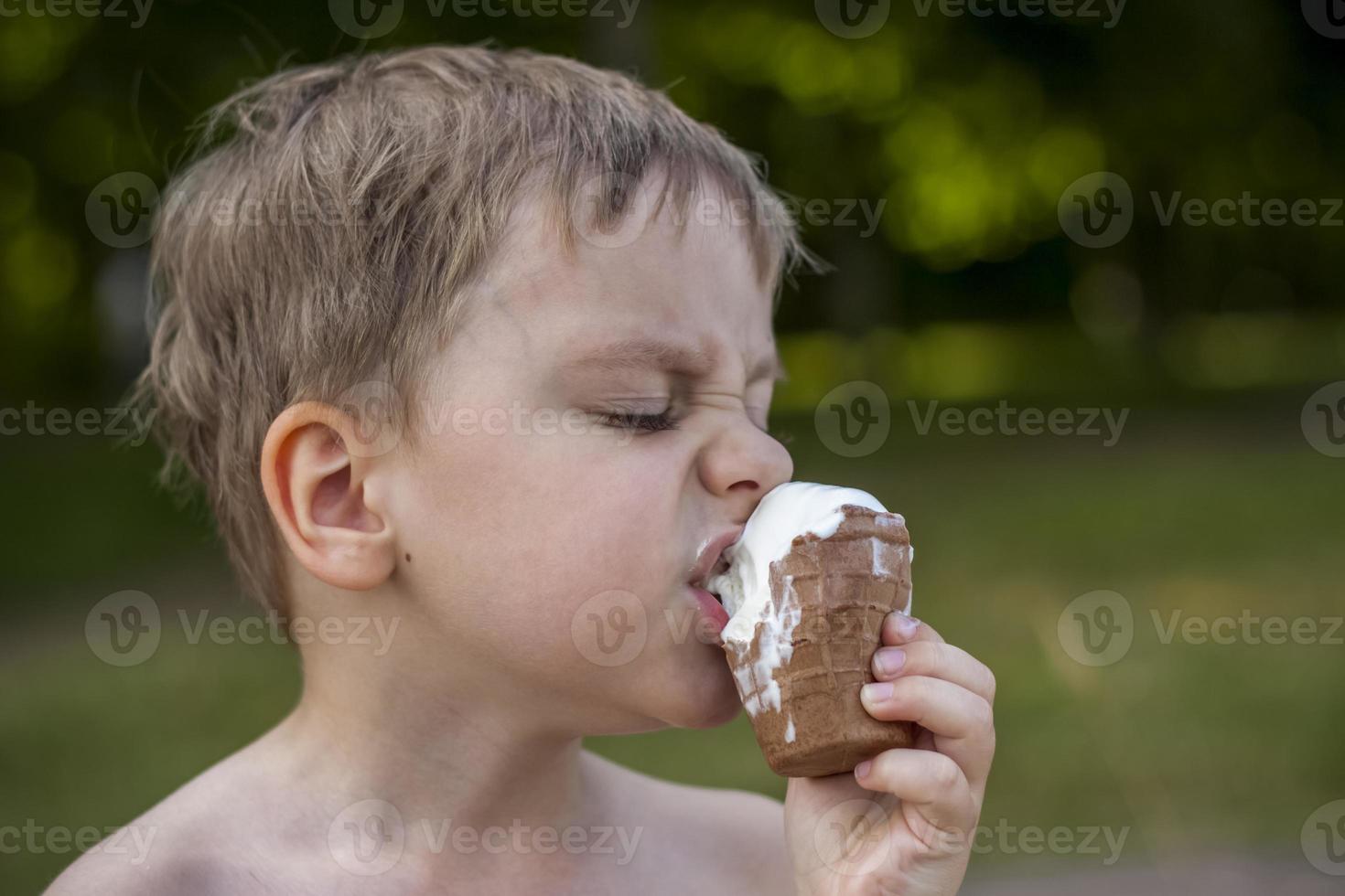 A cute blond boy appetizingly eats ice cream in the summer, sitting on the bank of the river. Cool off by the water. Funny facial expression. photo