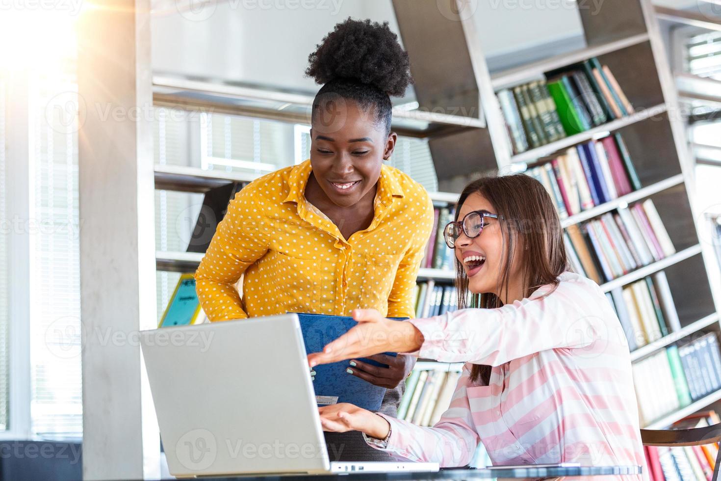 Young professionals work in company, using laptop and tablet. Two women are at table, talking with smiles, looking at screen of silvery pc and holding analytics in hands. photo