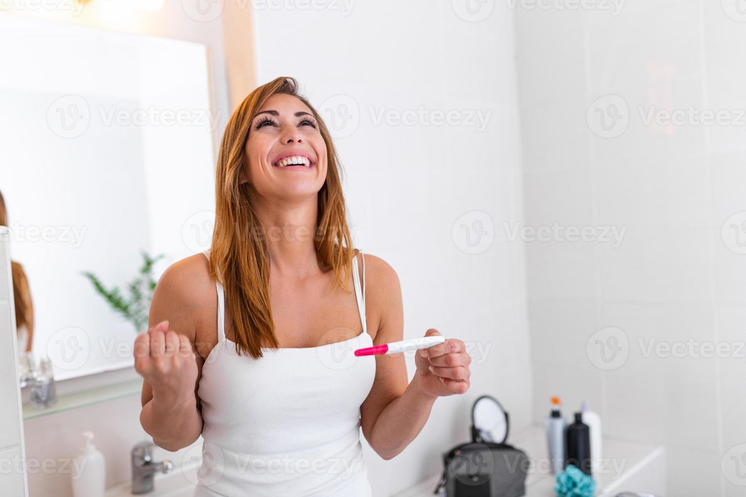 Close-up of young female dressed in white top, looking at pregnancy test result with positive smile, happy that she is going to have baby. Finally pregnant. photo