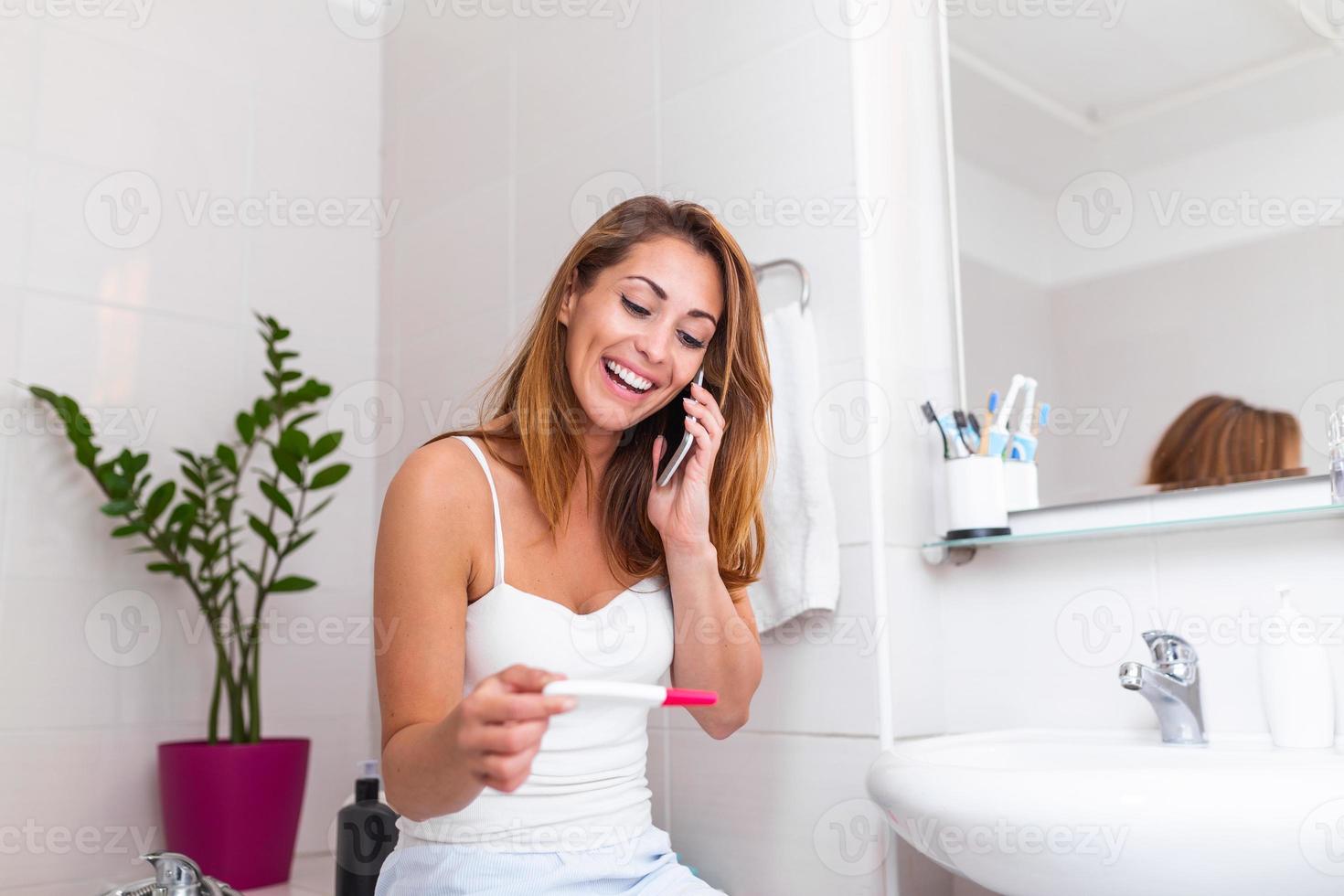 Smiling woman sitting in bathroom and holding positive pregnancy test and talking on her smart phone with the father to give him happy news. Smiling pregnant girl telling good news on mobile. photo