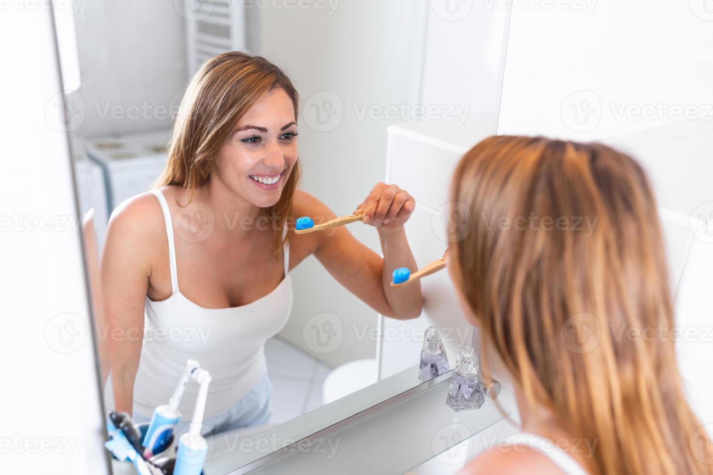 Pretty female brushing her teeth in front of mirror in the morning. Smiling young woman with healthy teeth holding a tooth brush photo