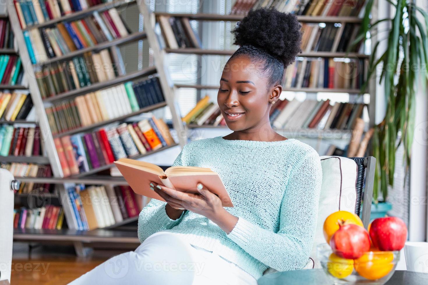 Young beautiful African American girl reading a book on the couch with the library bookshelves in the back. Beautiful woman on a white sofa reading a book photo