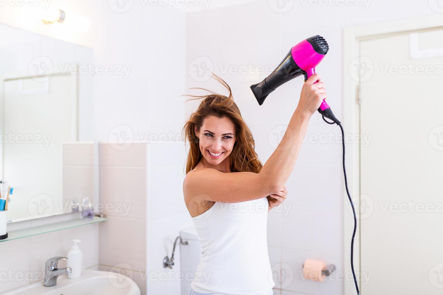 Beautiful girl using a hair dryer and smiling while looking at the mirror. Smiling woman drying hair with hair dry machine. Happy girl looking at mirror while using hair dryer in the bathroom. photo