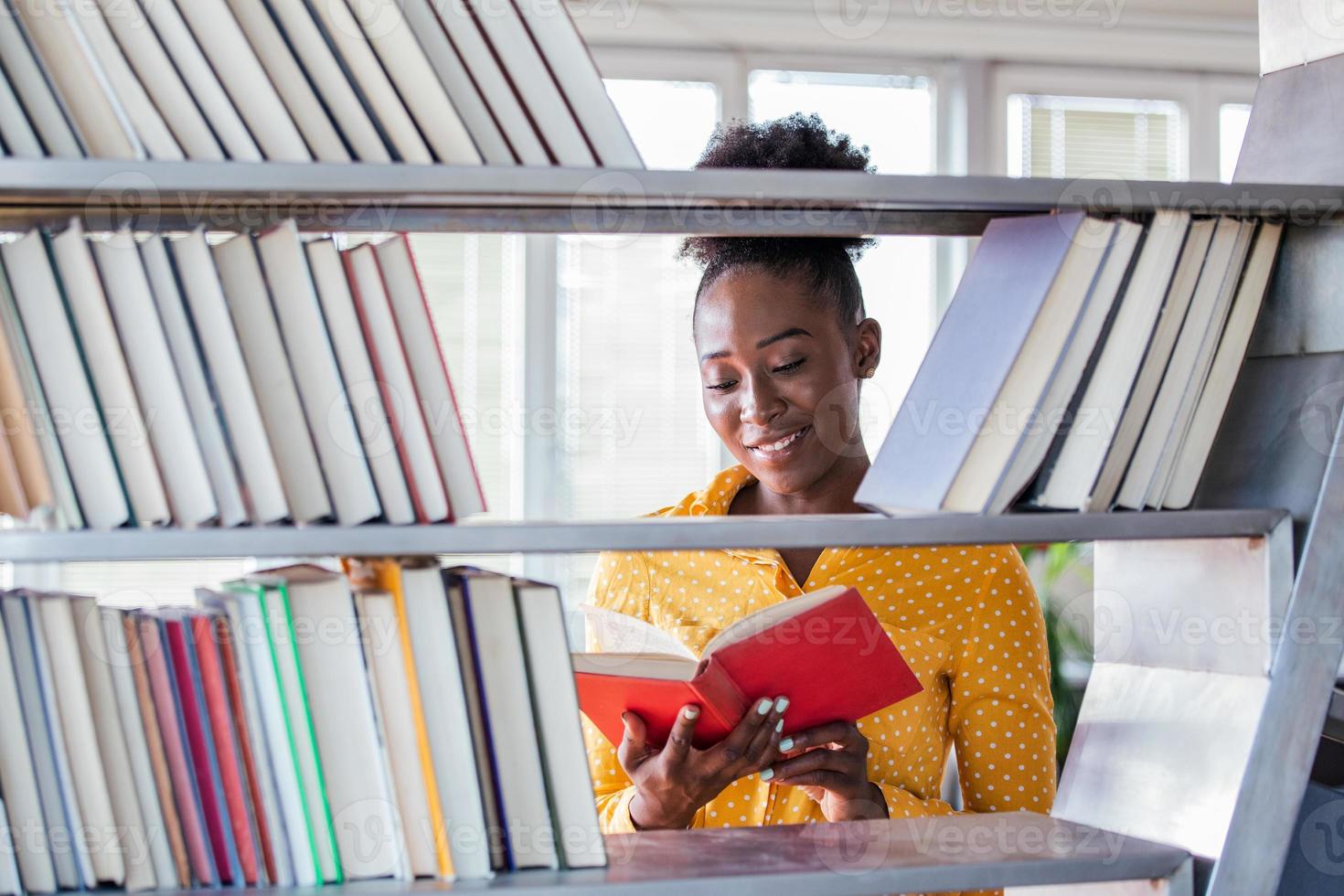 mujer estudiante universitaria afroamericana trabajando en la biblioteca sosteniendo un libro en las manos, luciendo inteligente. estanterías en la biblioteca. foto