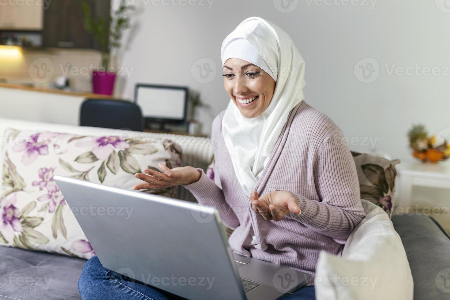 joven musulmana que tiene una videollamada a través de una computadora portátil en casa. feliz sonriente mujer musulmana sentada en un sofá, sofá y usando una laptop en la sala de estar en casa, aprendiendo idiomas, videollamadas. foto