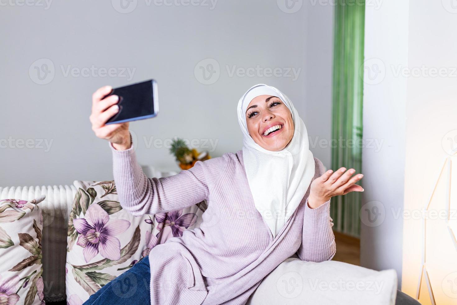 Young Muslim woman having video call via smart mobile phone at home. Happy smiling Muslim woman sitting on sofa, couch and using smart phone at living room at home, learning language, video calling. photo