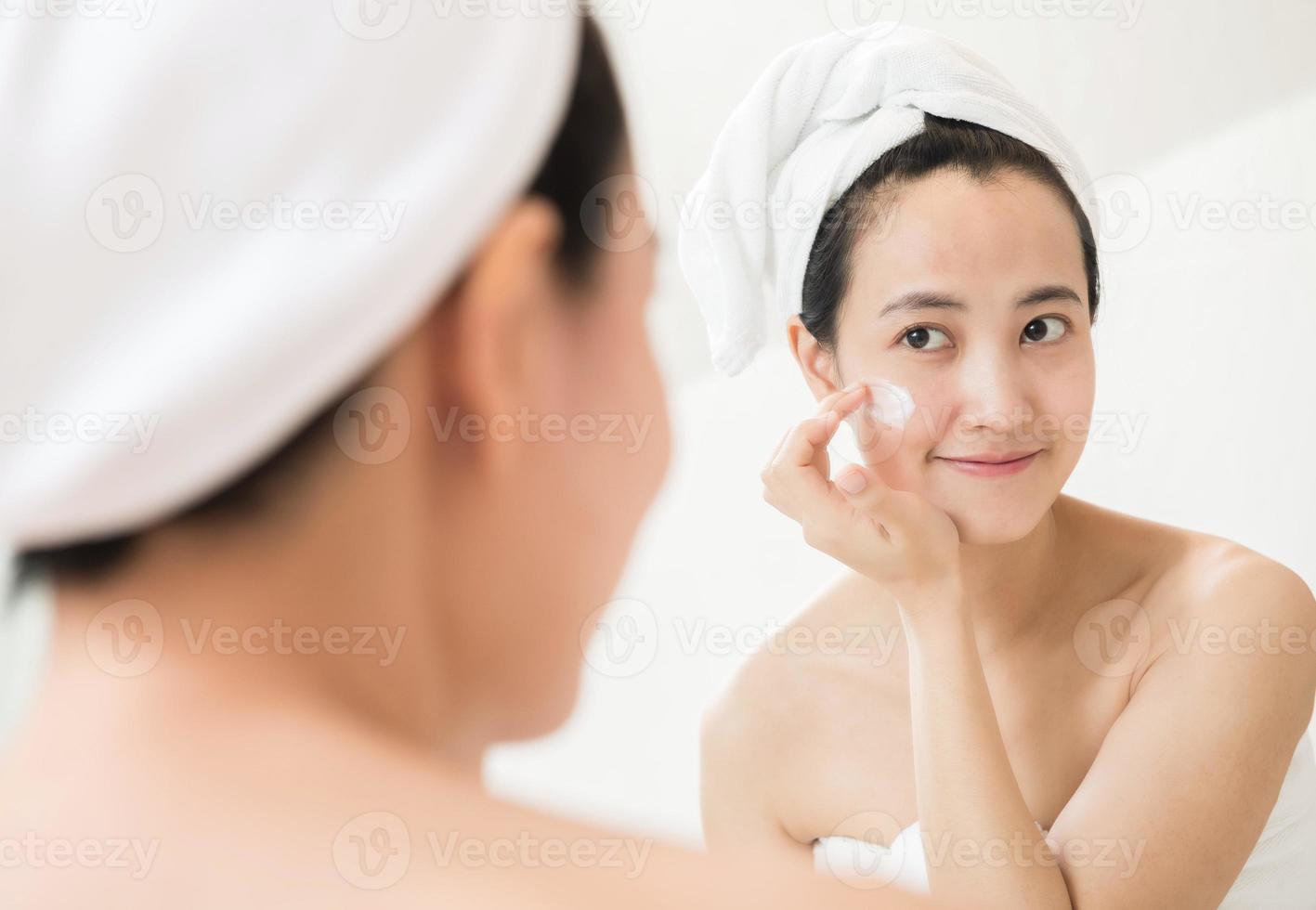 Happy young Asian woman applying face lotions while wearing a towel and touching her face in bathroom photo