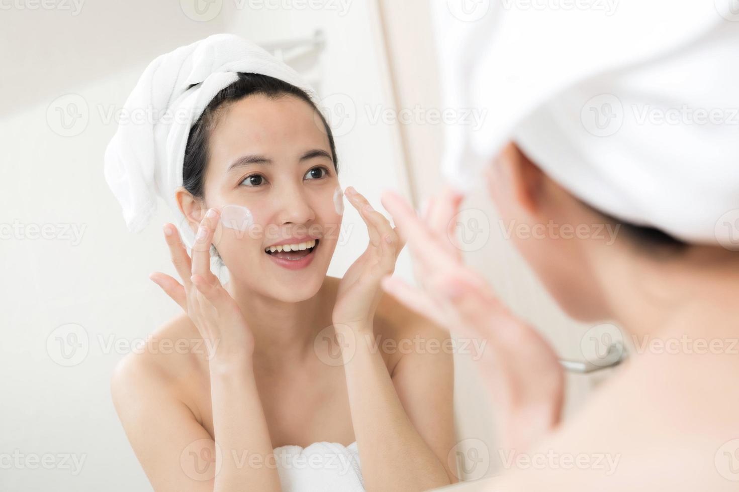 feliz joven asiática aplicando lociones faciales mientras usa una toalla y toca su cara en el baño foto