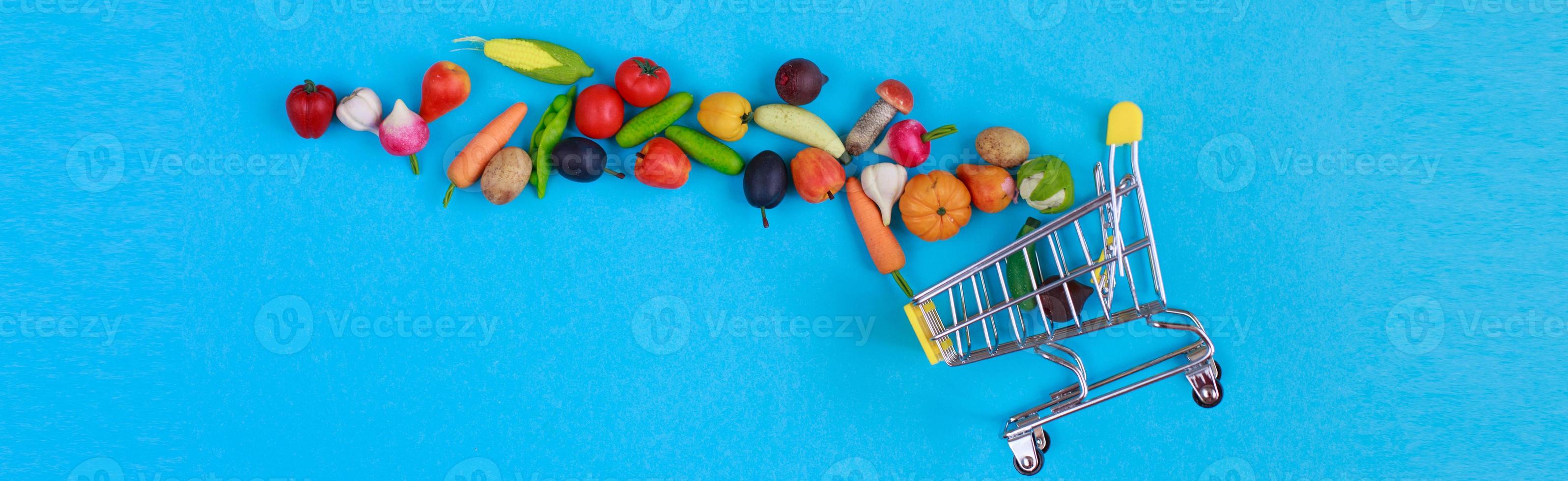 Metal shopping cart with fruits and vegetables on a blue background. Toy miniature shopping trolley photo