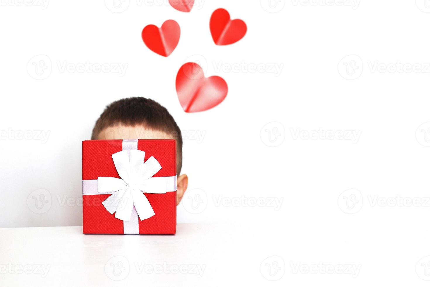 a young man holding a present box with red hearts on a white background. child covers his face with a box photo