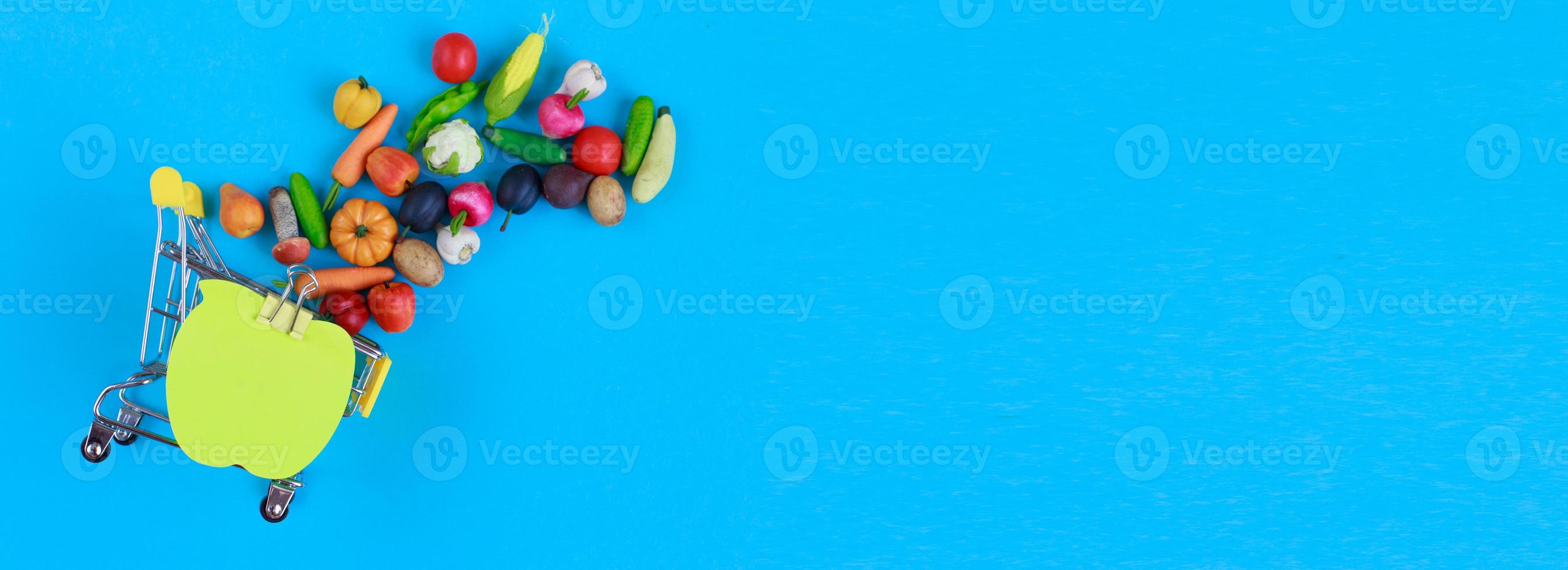 Metal shopping cart with fruits and vegetables on a blue background. Toy miniature shopping trolley photo