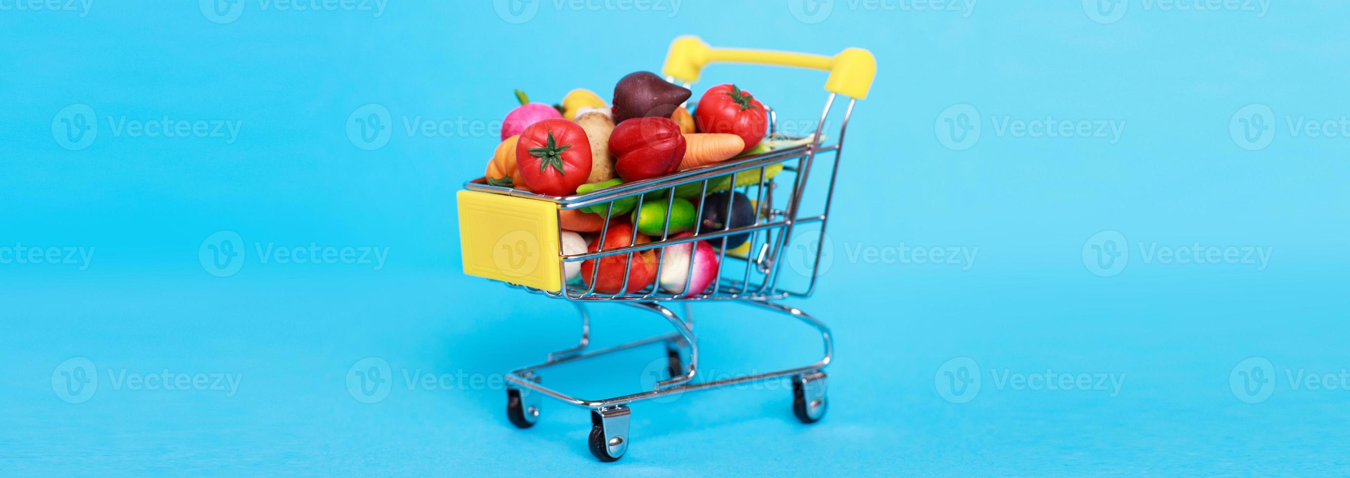 Metal shopping cart with fruits and vegetables on a blue background. Toy miniature shopping trolley photo