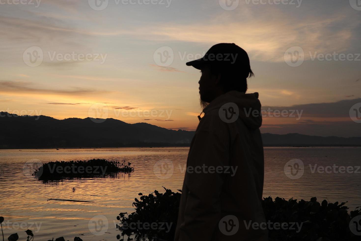 silueta de un joven parado junto al lago disfrutando de la puesta de sol. ambiente tranquilo en la naturaleza foto