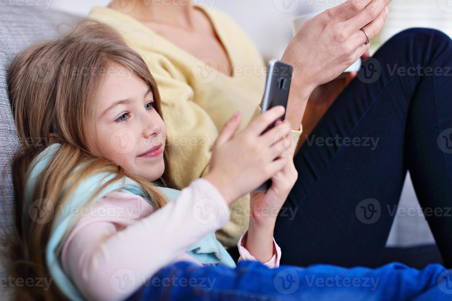 Young mom and her little daughter using smartphone and sitting on sofa at home photo